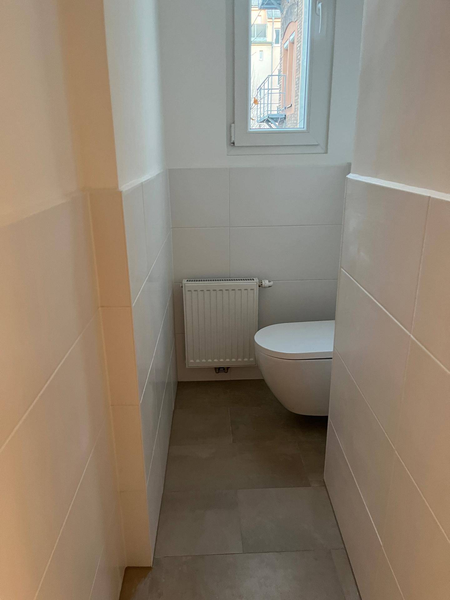 Helles Gäste-WC mit wandhängender Toilette, Fenster und Heizkörper in neutralen Farbtönen.