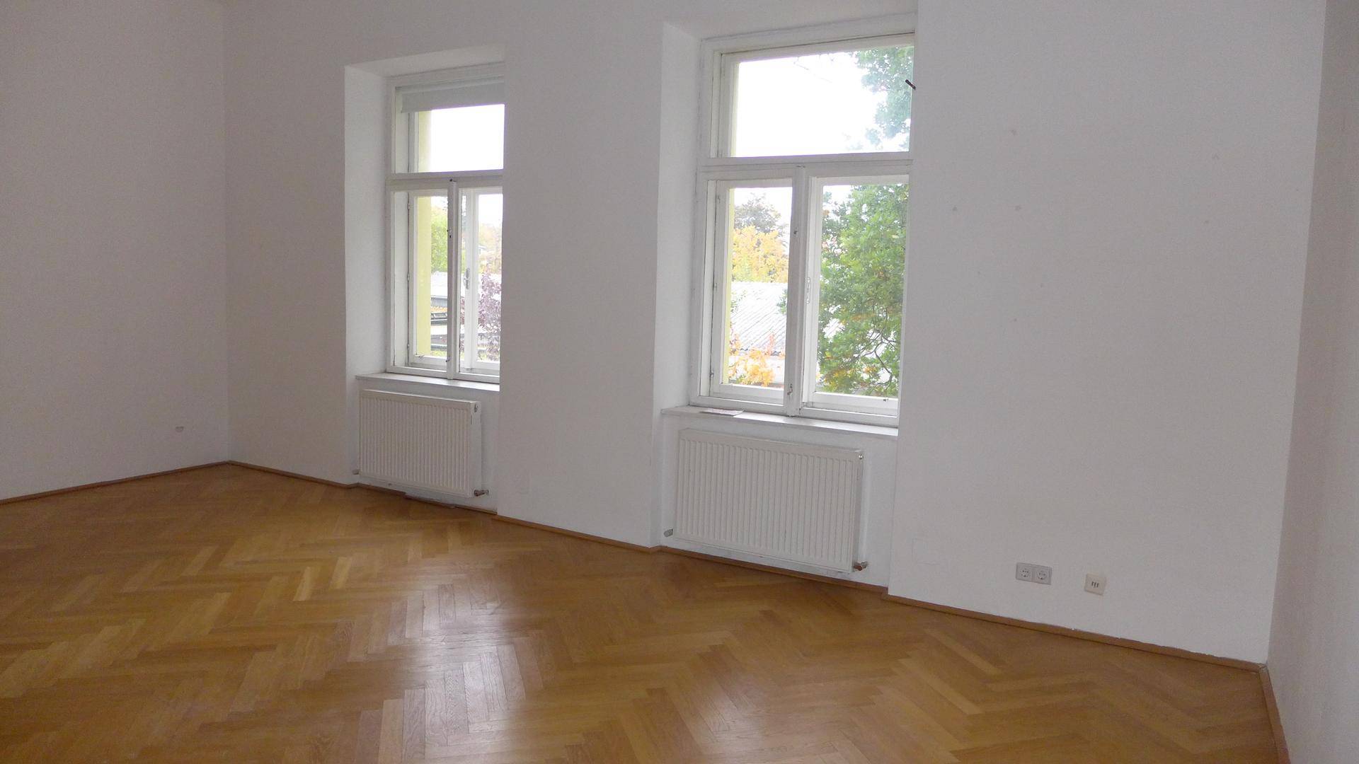 Geräumiges Wohnzimmer mit Fischgrätparkettboden und zwei großen Fenstern für viel Tageslicht.