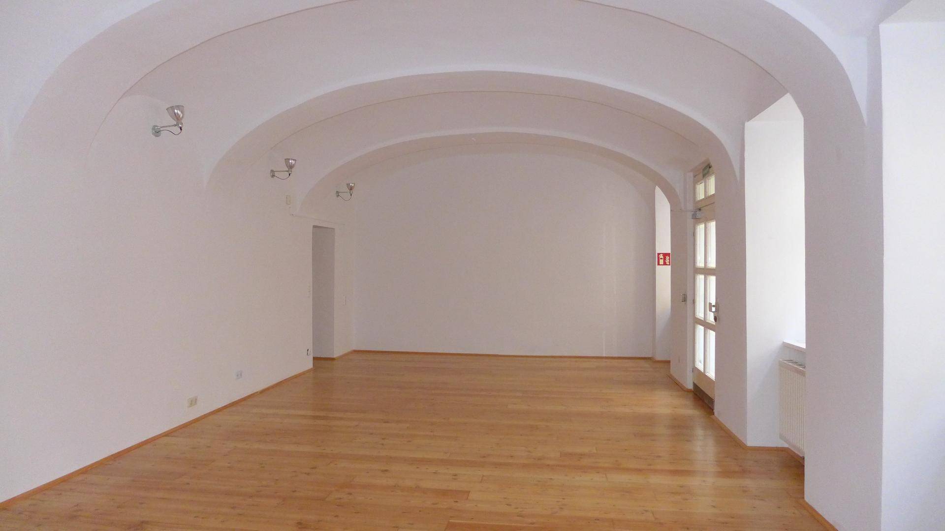 Geräumiger, leerer Raum mit beeindruckenden Gewölbedecken und hellem Holzfußboden, gut beleuchtet.