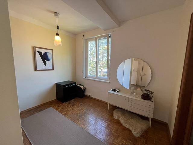 Kleines Zimmer mit Parkettboden, Kommode, rundem Spiegel und Fenster.