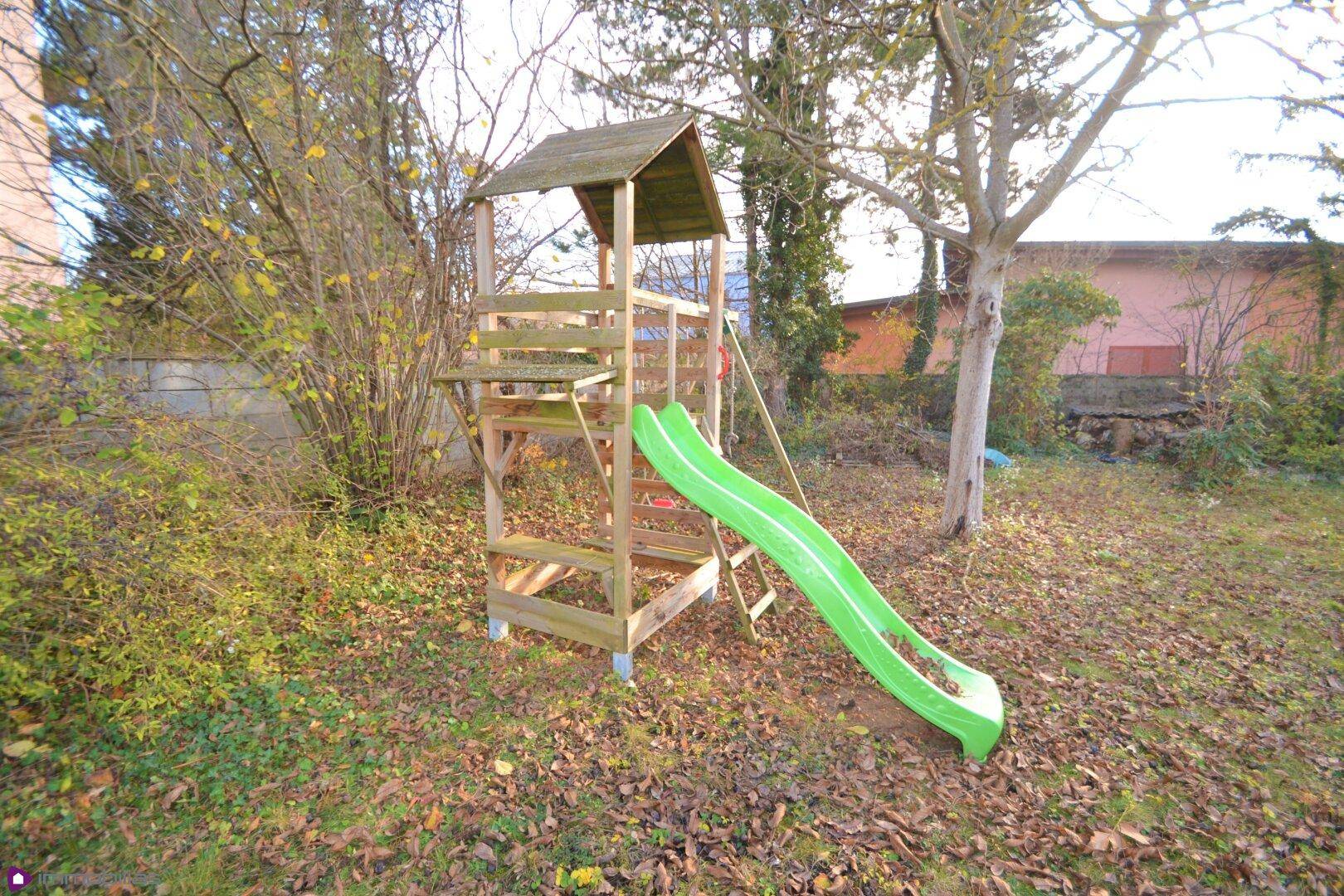 Gartenbereich mit einem Holzspielturm und Rutsche, umgeben von Bäumen und Sträuchern.