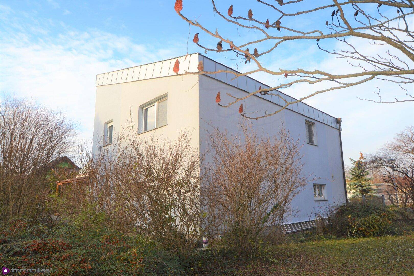 Moderne Hausfassade mit weißem Putz und schrägen Dachlinien, umgeben von Vegetation.