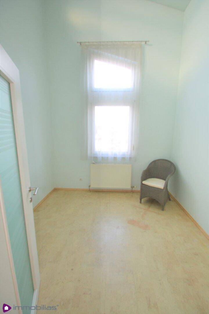 Helles Zimmer mit hellem Boden, Fenster und einem Korbstuhl, ideal als Büro oder Gästezimmer.