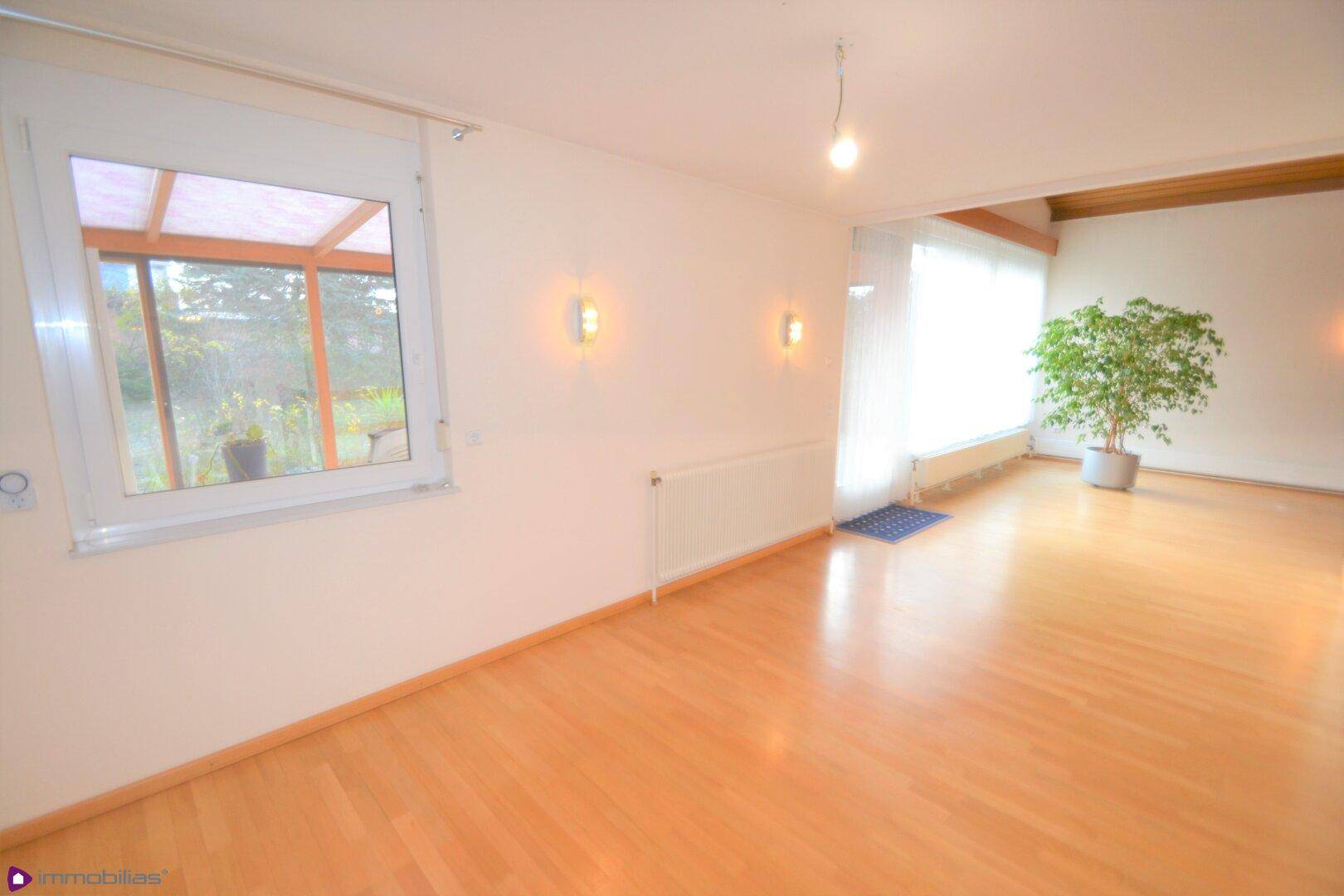 Helles Wohnzimmer mit Parkettboden und einem Fenster, das den Blick auf den Wintergarten freigibt.
