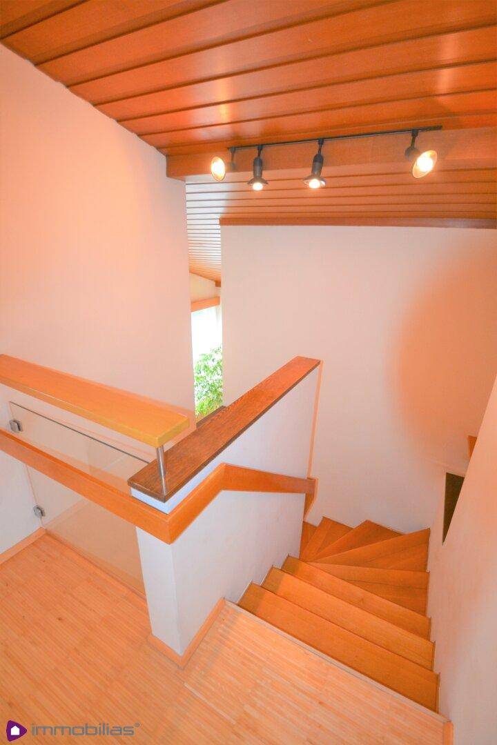 Holztreppe mit Glasgeländer führt in die obere Etage, beleuchtet von Deckenspots.