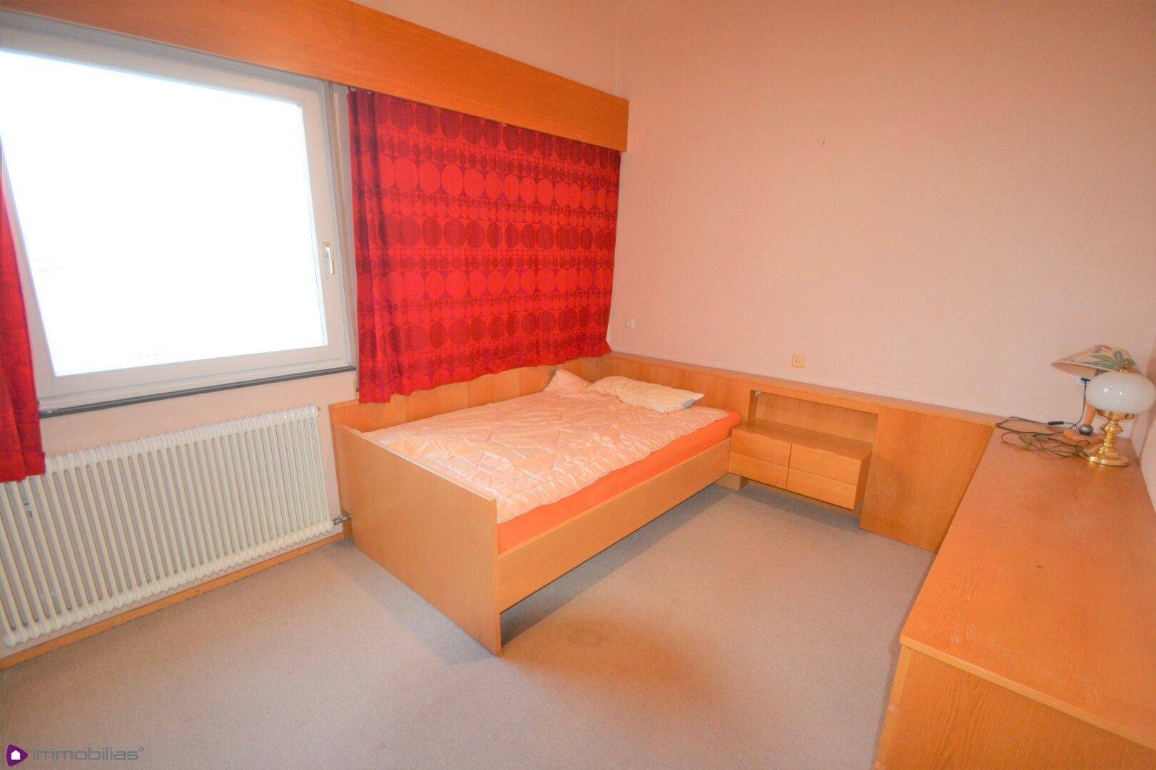 Schlafzimmer mit Teppichboden, Holzbett und integrierten Möbeln unter dem Fenster.