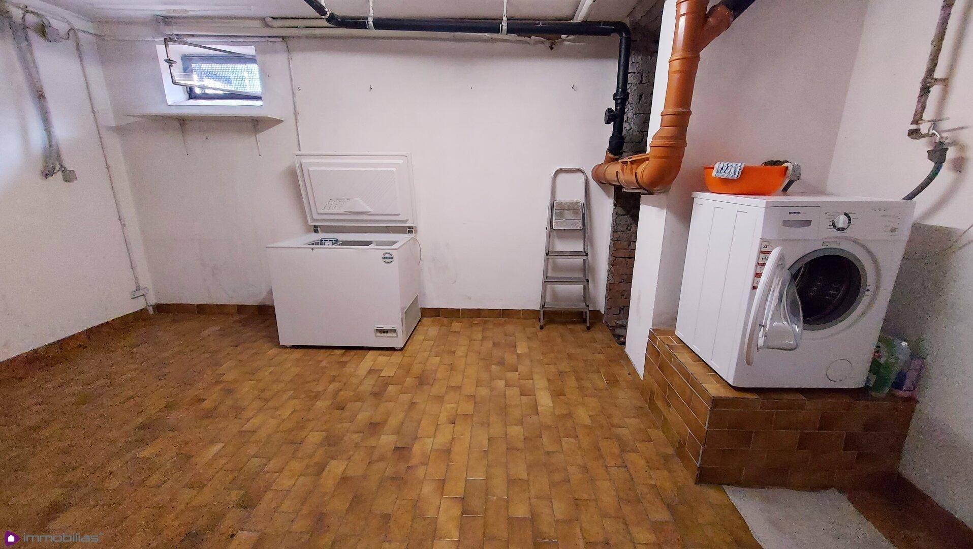 Waschküche im Keller mit Waschmaschine, Gefriertruhe und sichtbaren Rohren.