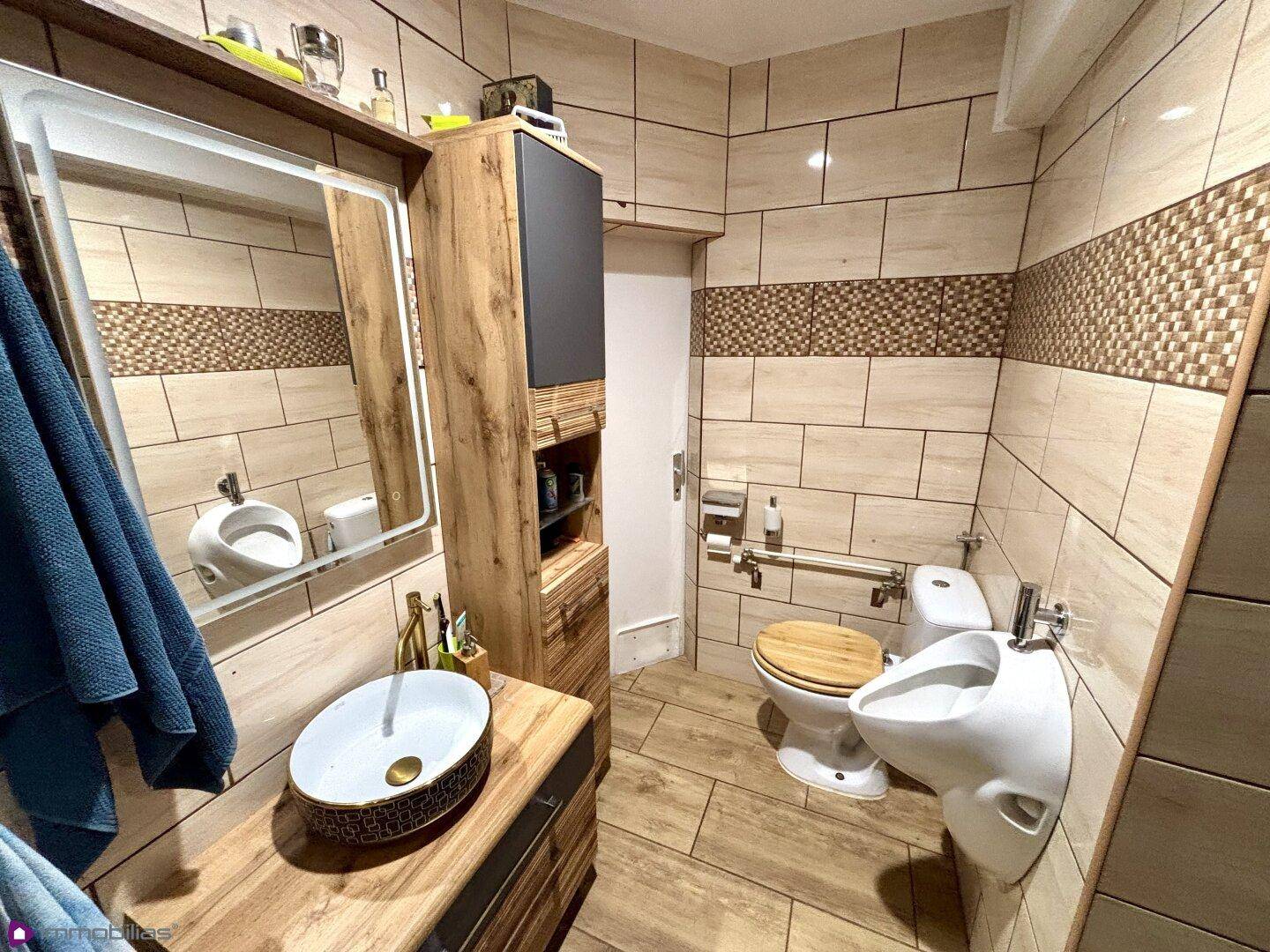 Modernes Badezimmer mit beigen Fliesen, Holzwaschtisch, Bidet und Haltegriffen.