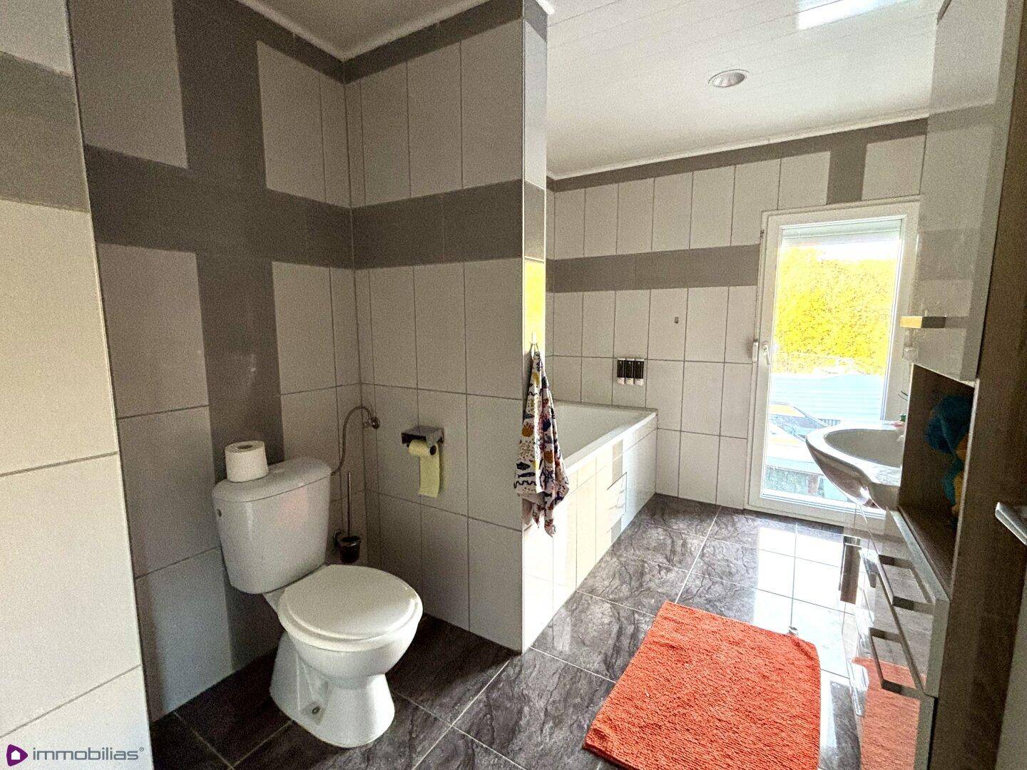 Badezimmer mit grauen und weißen Fliesen, Badewanne, Toilette und Fenster.