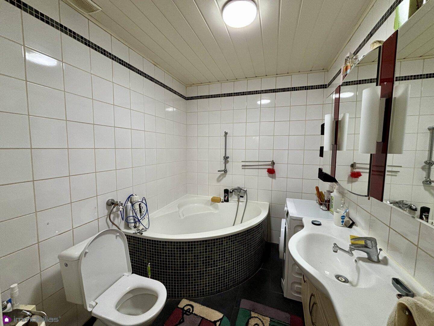 Badezimmer mit weißen Fliesen, Eckbadewanne, Toilette und Waschbecken.