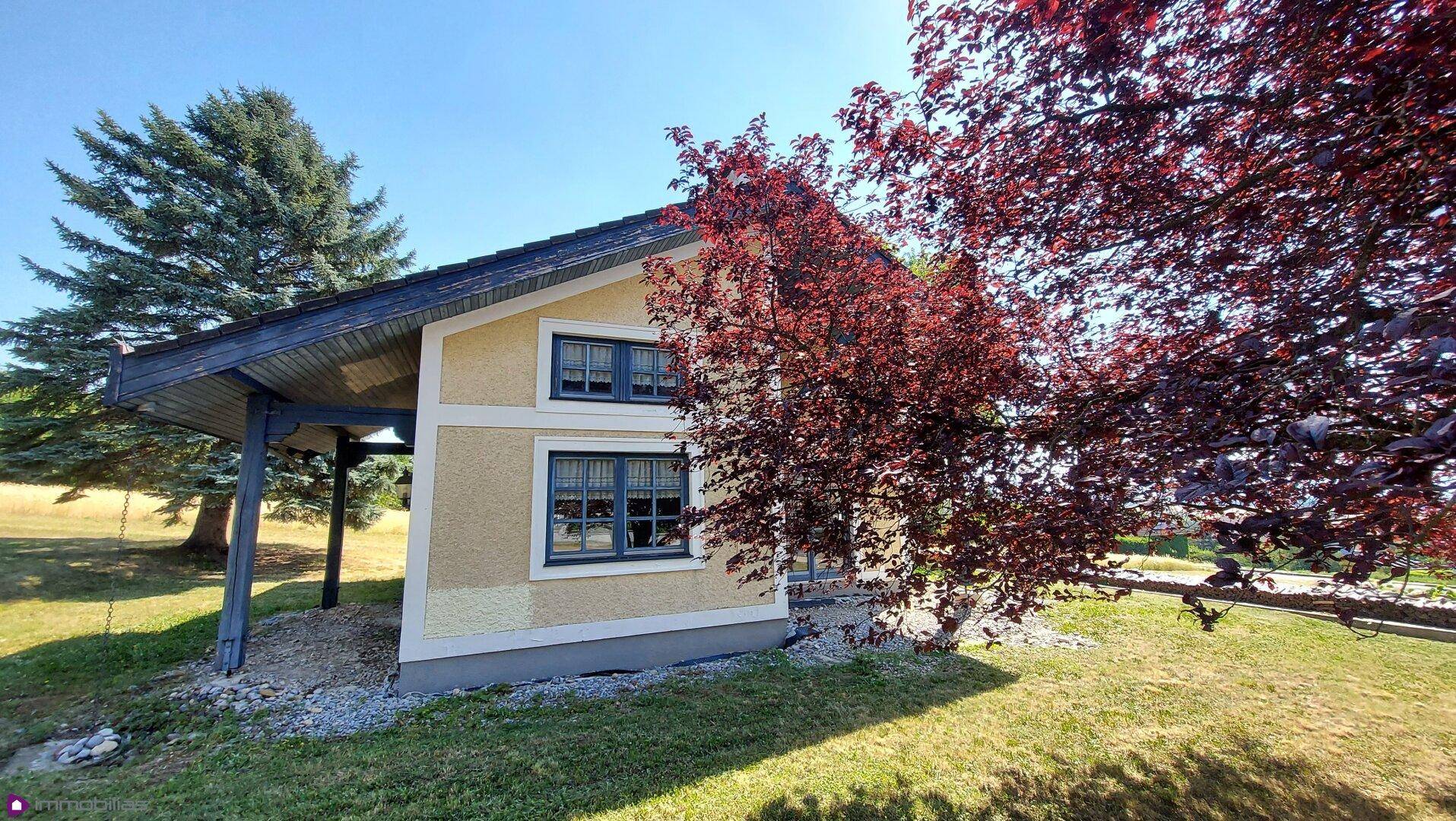 Seitenansicht des Hauses mit gelber Fassade, umgeben von Grün und einem großen Baum.