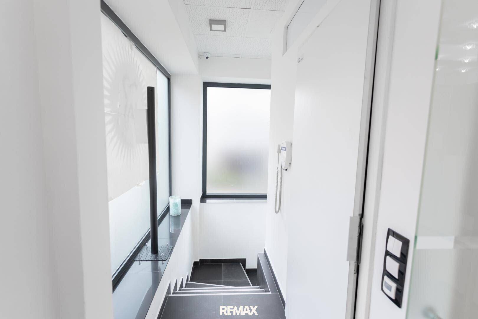 Heller Treppenaufgang mit weißen Wänden, dunklen Stufen, einem Fenster mit Milchglasfolie und Gegensprechanlage.