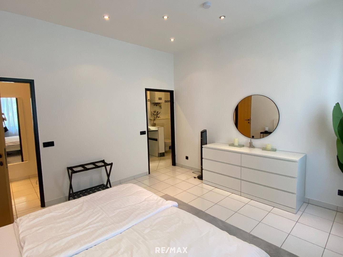 Schlafzimmer mit Kommode und rundem Spiegel, bietet Zugang zu einem weiteren Raum.