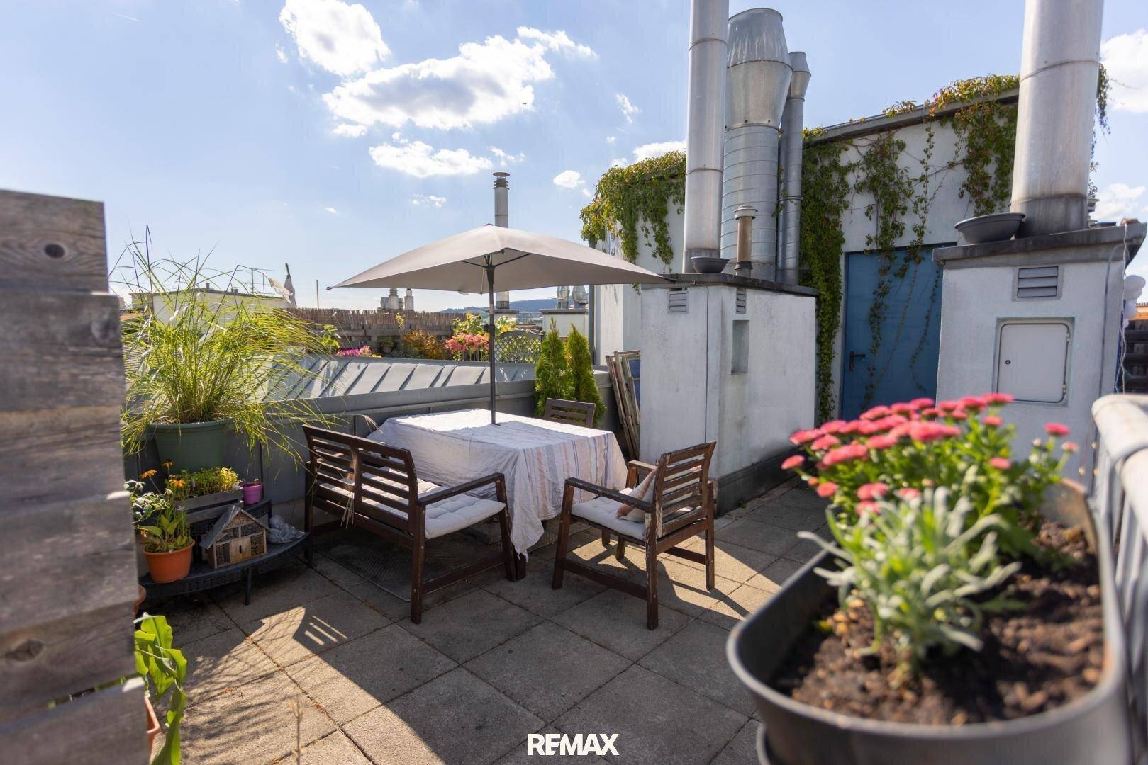 Gemütliche Dachterrasse mit Esstisch, Stühlen und Sonnenschirm unter blauem Himmel.