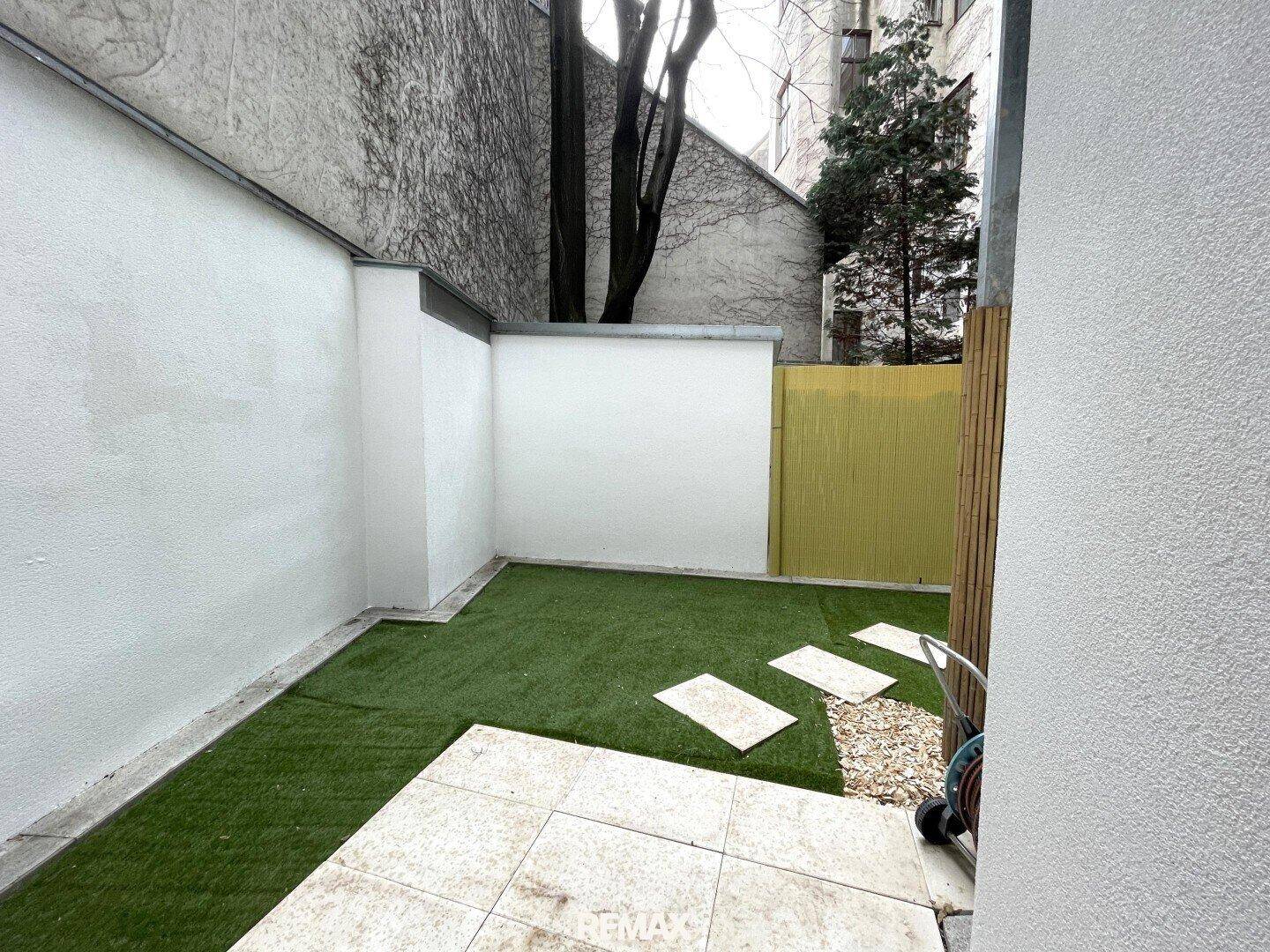 Seitlicher Blick auf den kleinen Garten mit Kunstrasen und Steinplatten in einem ummauerten Bereich.