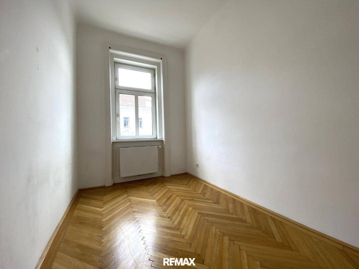 Helles Zimmer mit klassischem Fischgrätparkett, einem Fenster und Heizkörper.