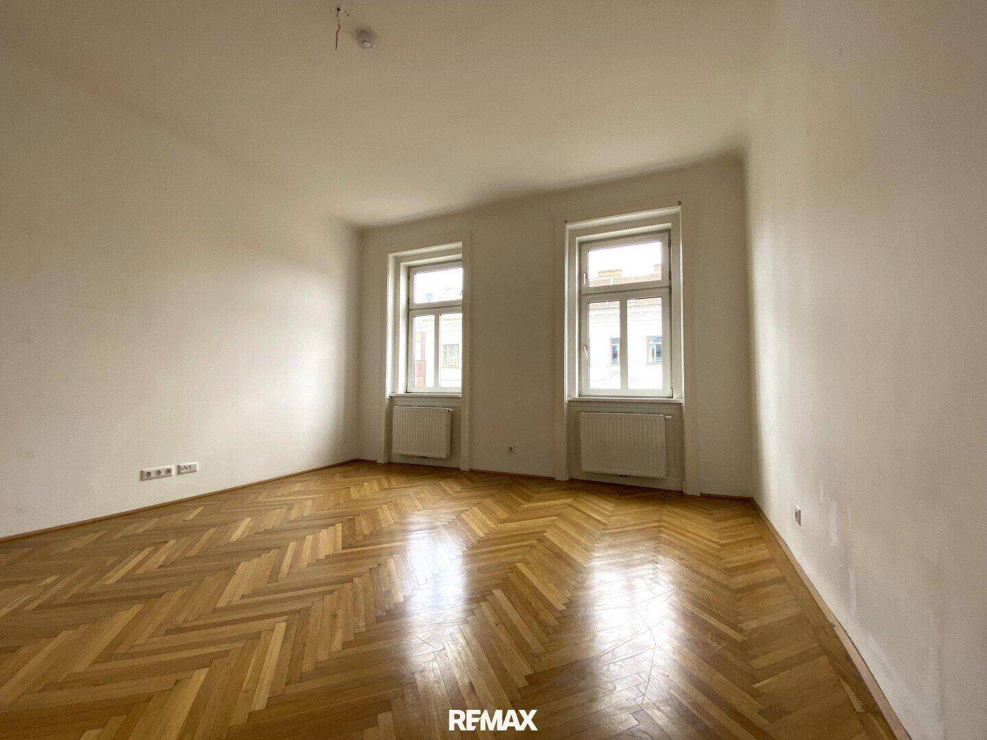 Geräumiges Zimmer mit klassischem Fischgrätparkett und viel Tageslicht durch zwei hohe Fenster.