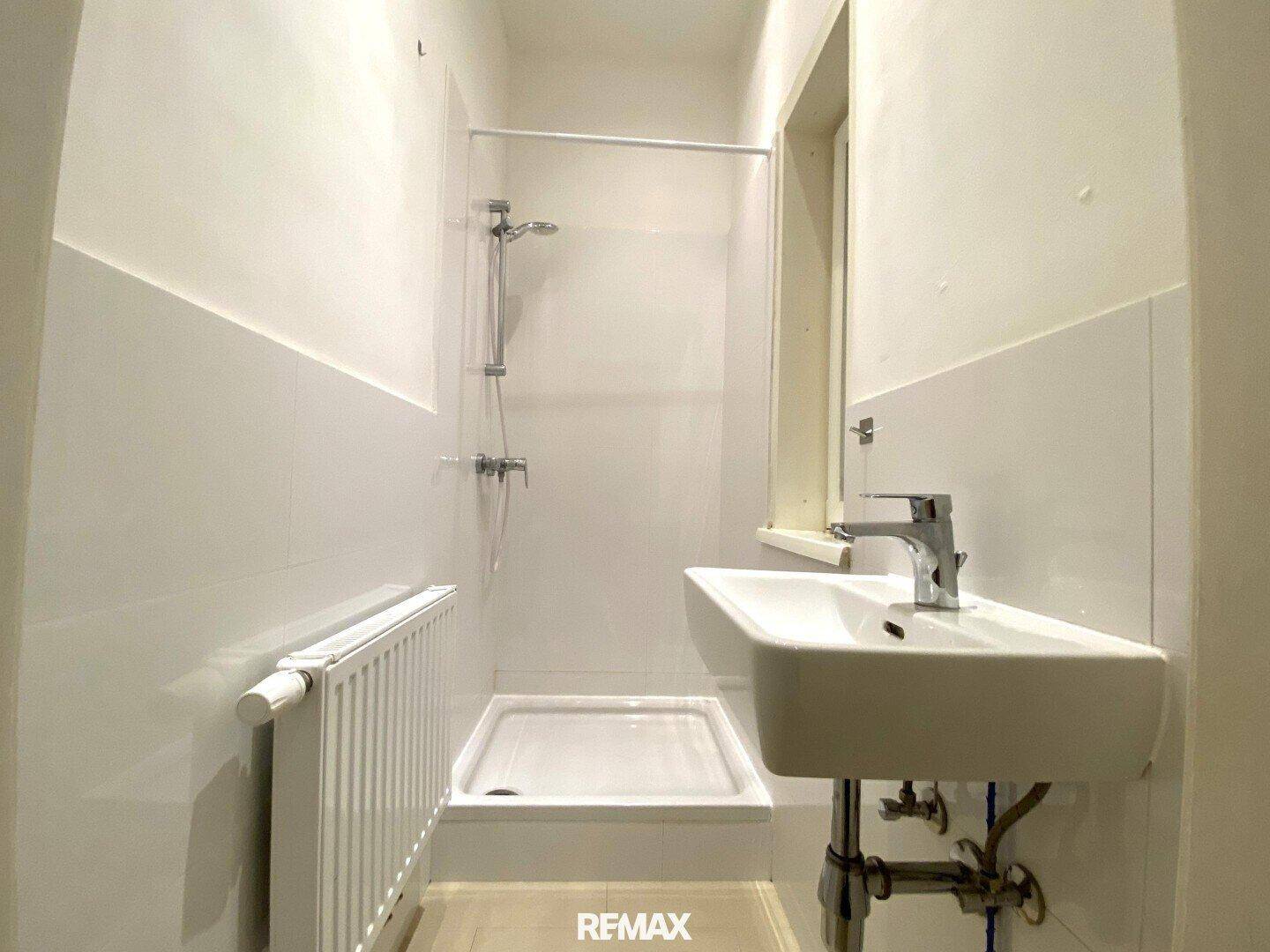 Helles Badezimmer mit Dusche, Waschbecken, Heizkörper und einem kleinen Fenster für natürliche Belüftung.