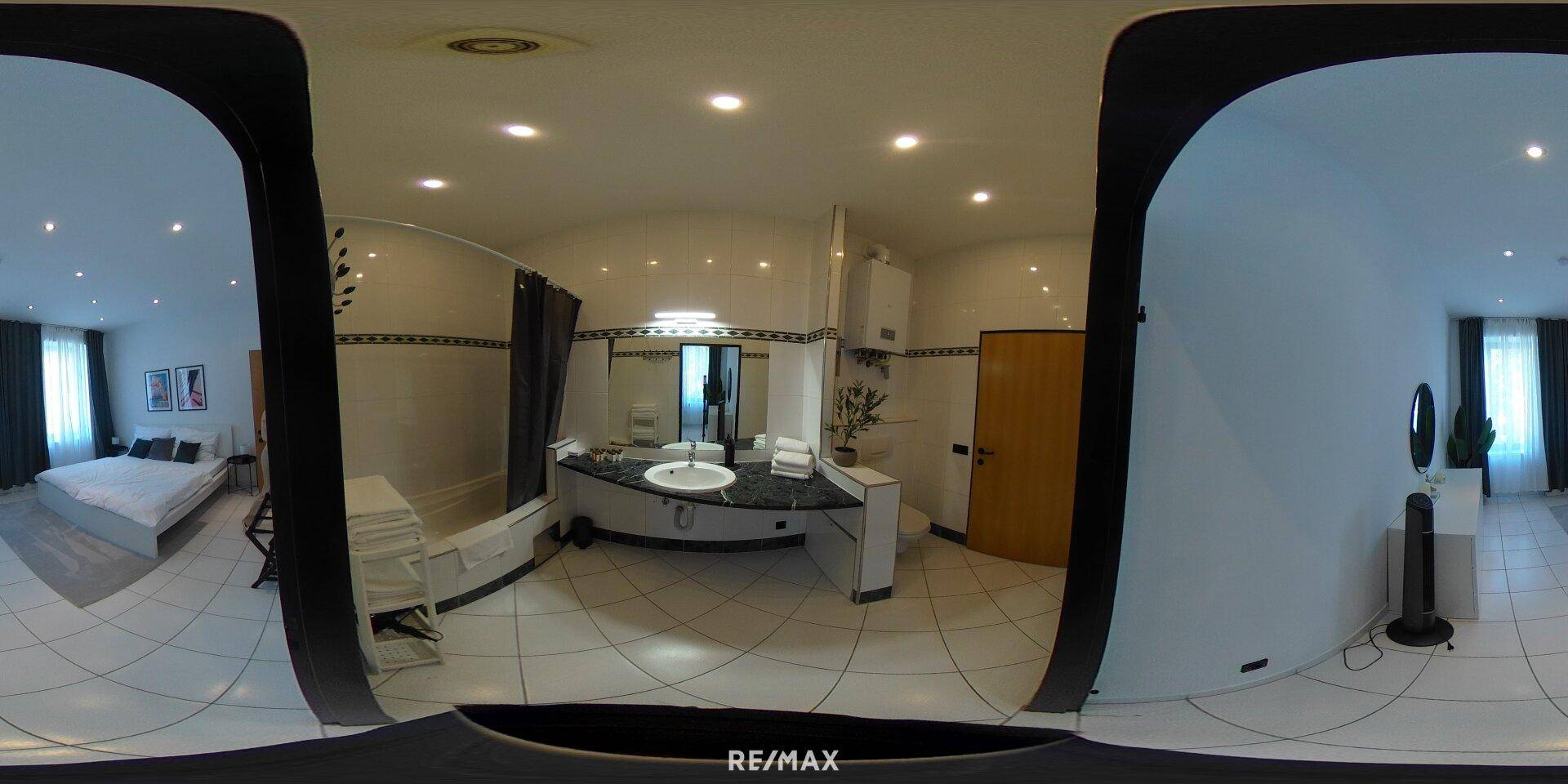Panoramablick auf das Badezimmer mit Badewanne und Waschtisch, mit Blick in ein Schlafzimmer.