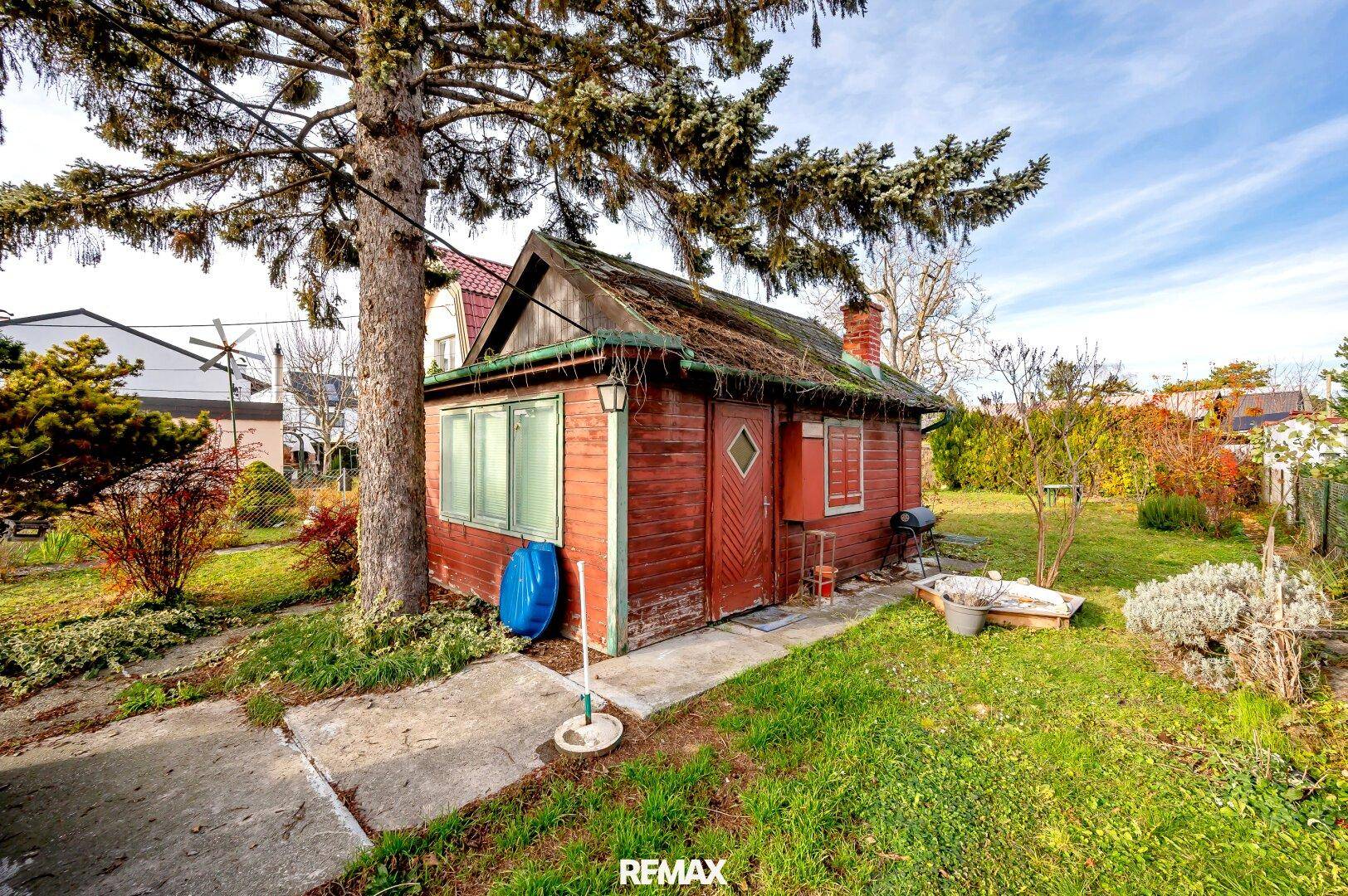 Kleines rotes Holzhaus im Garten unter einem großen Baum mit Grillbereich.