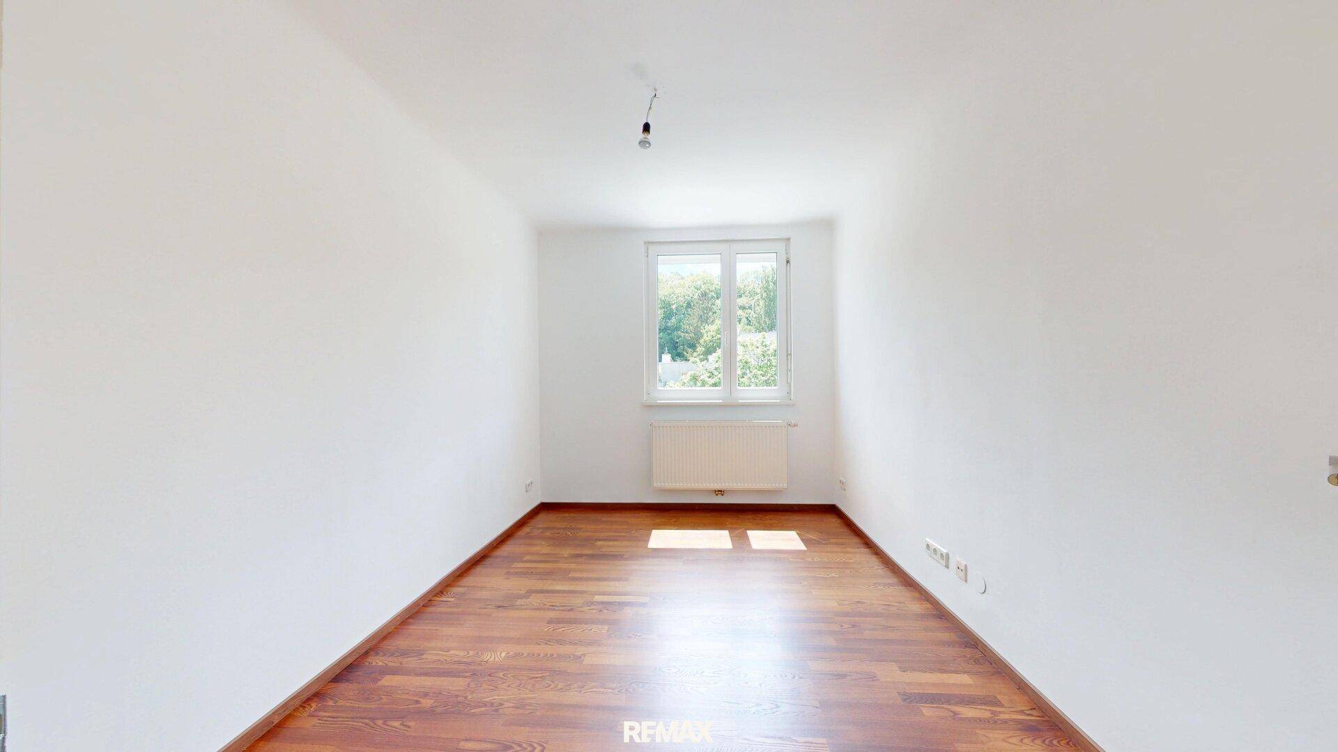 Helles, unmöbliertes Zimmer mit Laminatboden und einem Fenster mit Blick auf Bäume.
