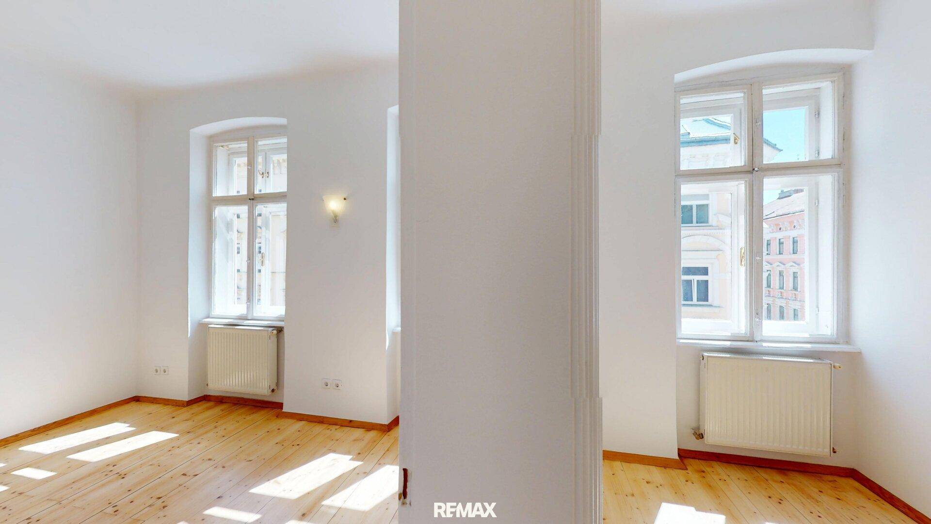Heller, leerer Raum mit Holzboden und großen Fenstern, die viel Tageslicht hereinlassen.