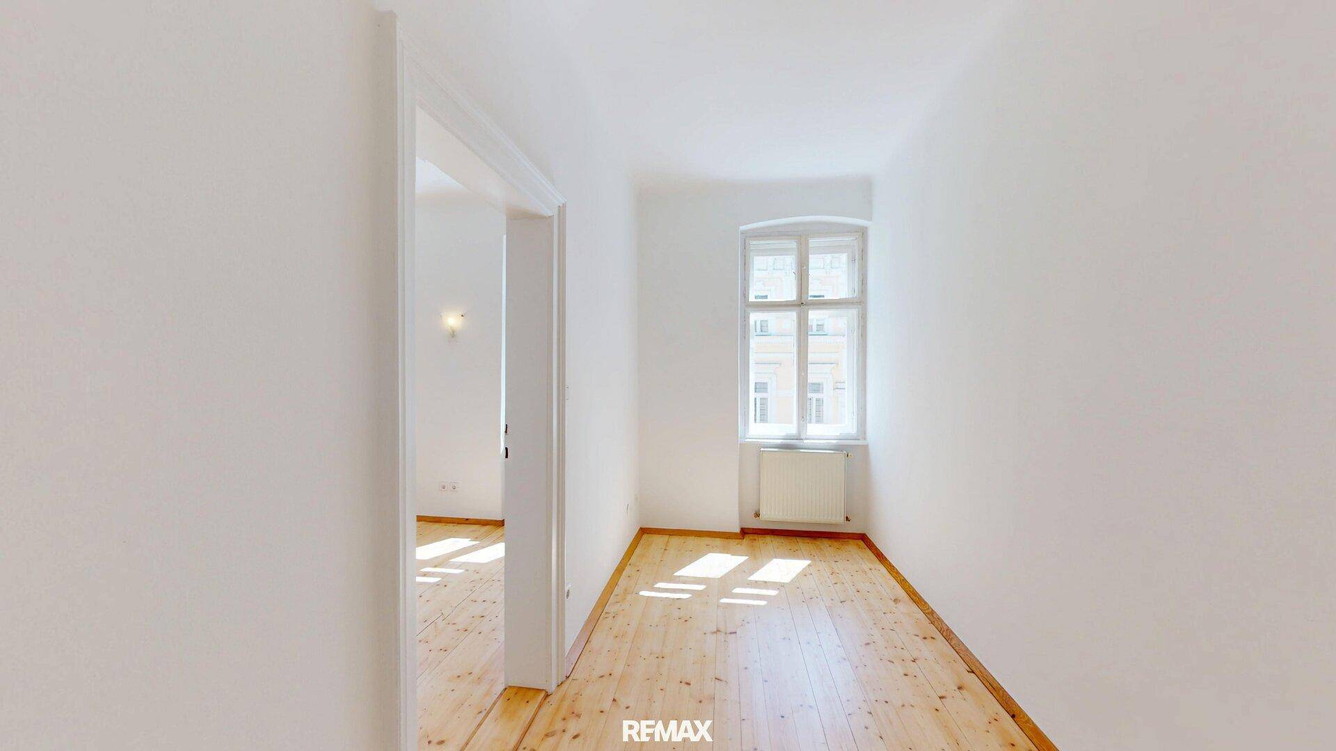 Schmaler Flur mit Holzboden und einem Fenster, der zu einem weiteren hellen Raum führt.