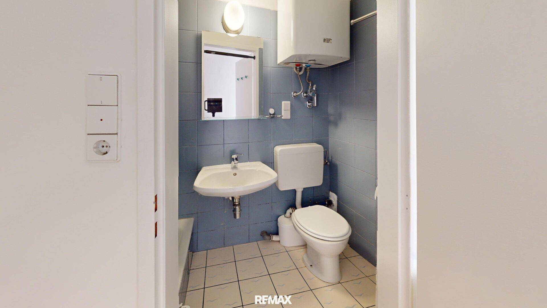 Kleines Badezimmer mit blauen Wandfliesen, WC, Waschbecken und einem Warmwasserboiler, funktional eingerichtet.