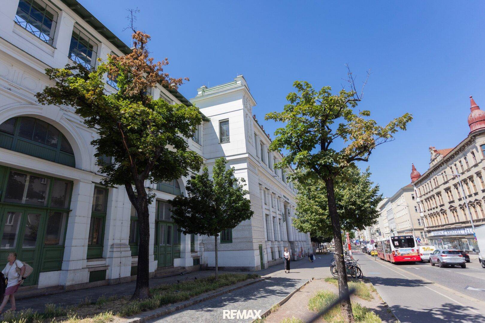 Stadtansicht mit einem großen weißen Gebäude, Bäumen und einem Bus auf der Straße unter strahlendem Sonnenschein.