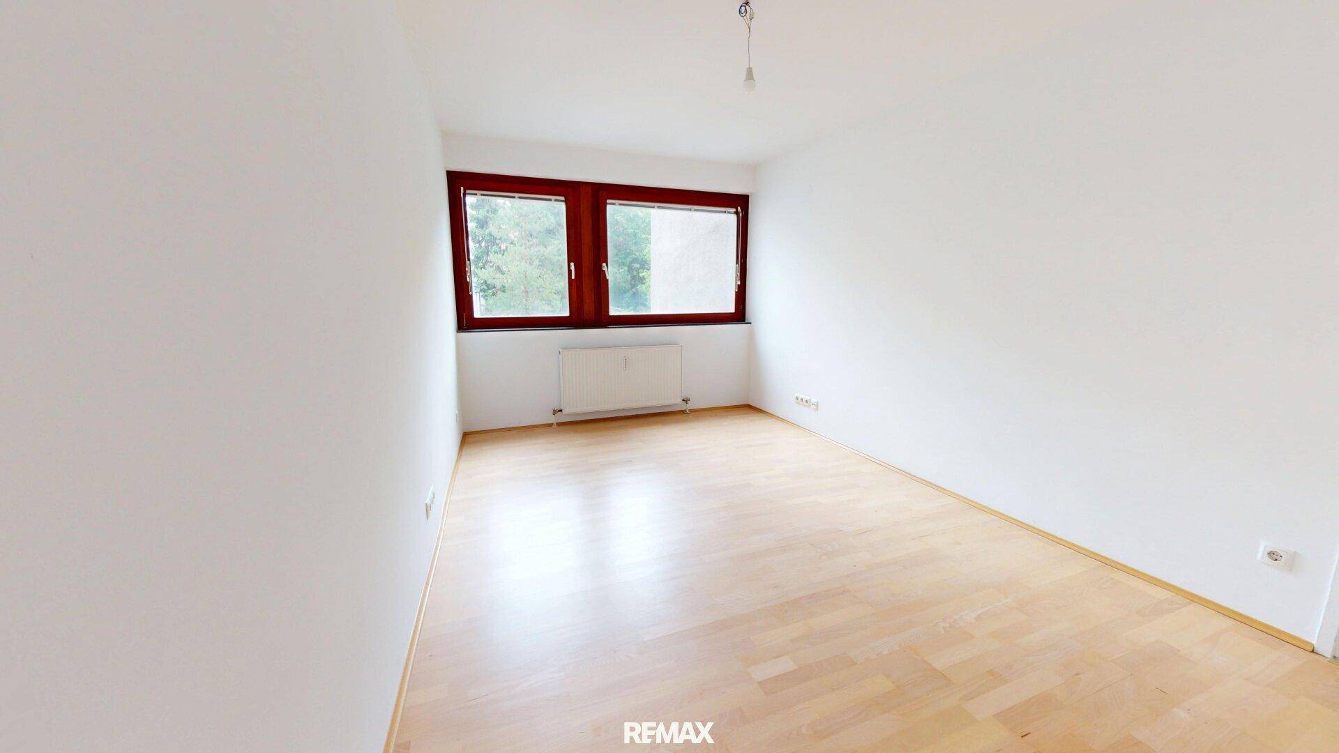 Leeres Schlafzimmer mit hellem Parkettboden, weißen Wänden und einem Fenster mit Blick ins Grüne.