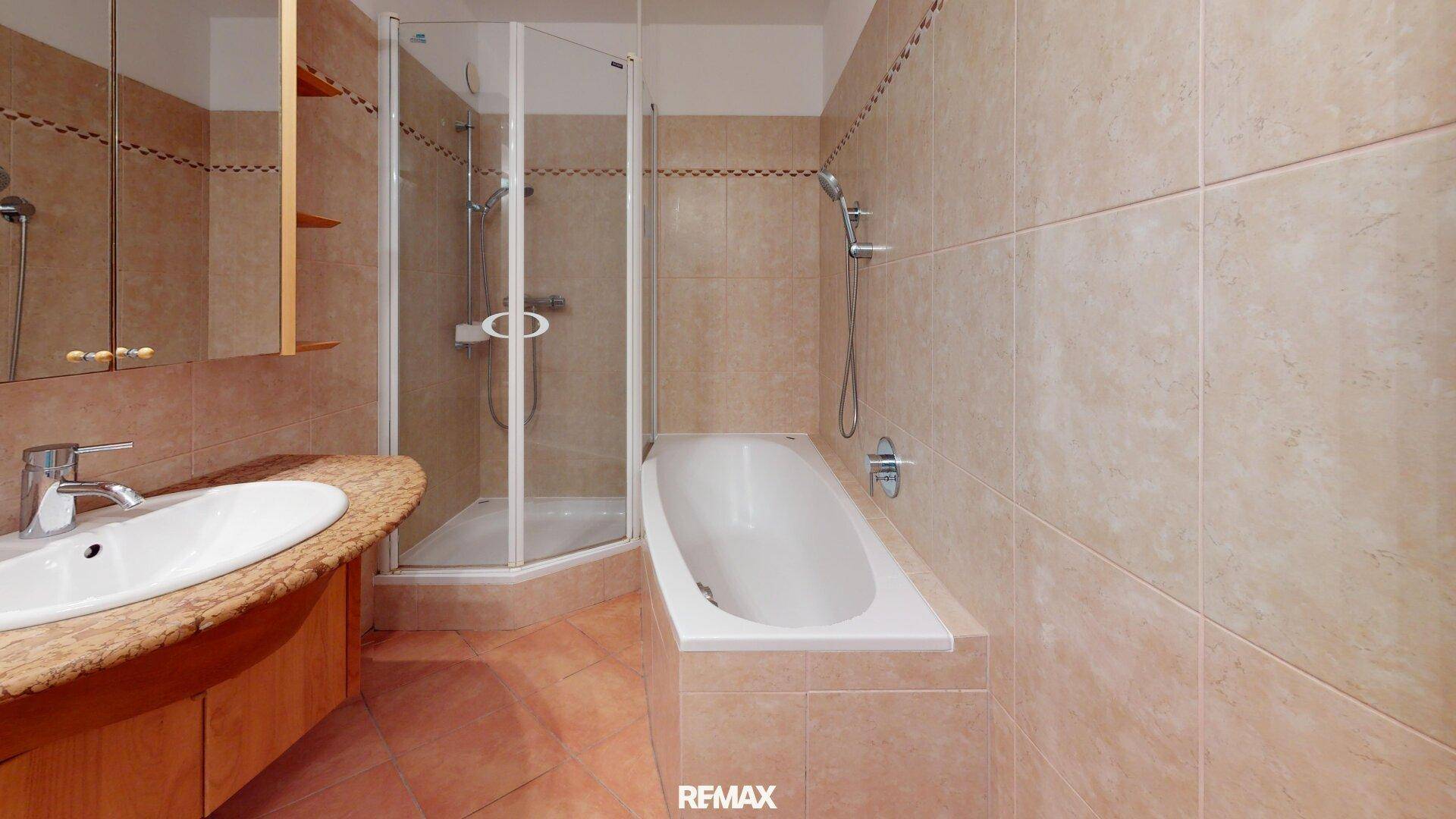 Badezimmer mit beigefarbenen Fliesen, Badewanne, Eckdusche und einem Waschbecken mit Unterschrank.