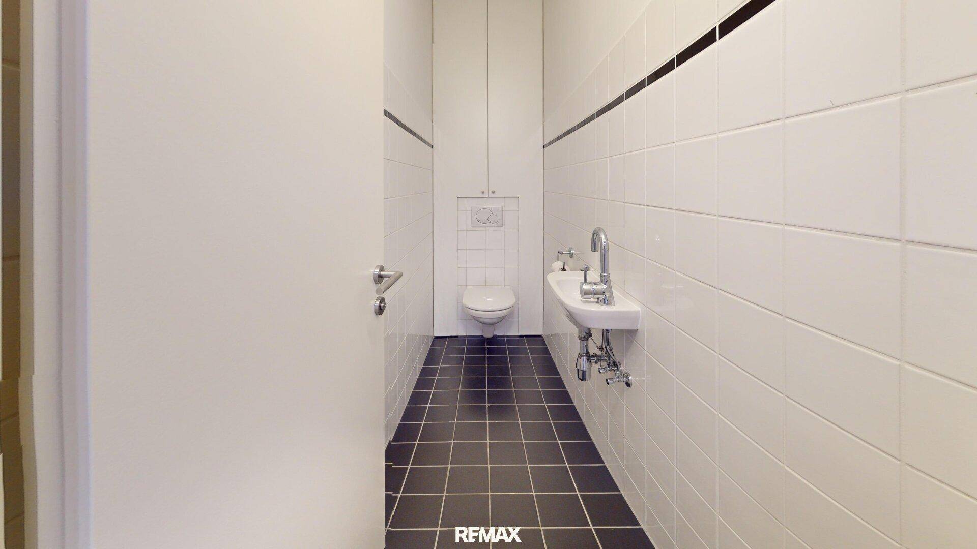 Separates WC mit weißen Wandfliesen, dunklem Boden und einem kleinen Handwaschbecken.