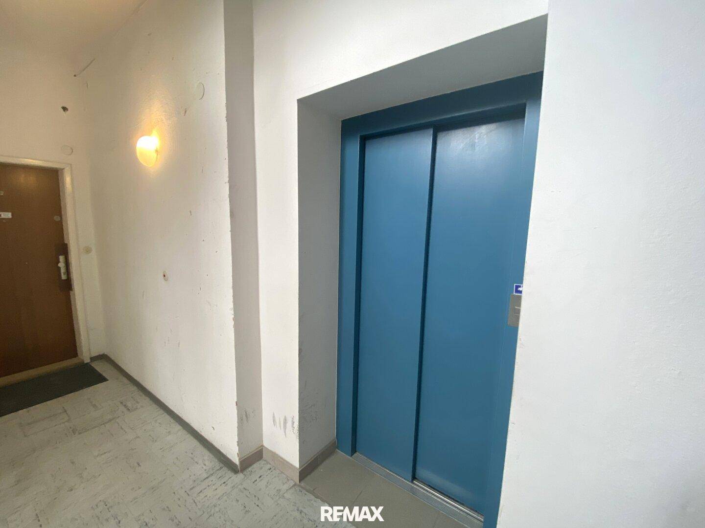 Schlichter Flur mit blauem Aufzug und einer Wohnungstür.