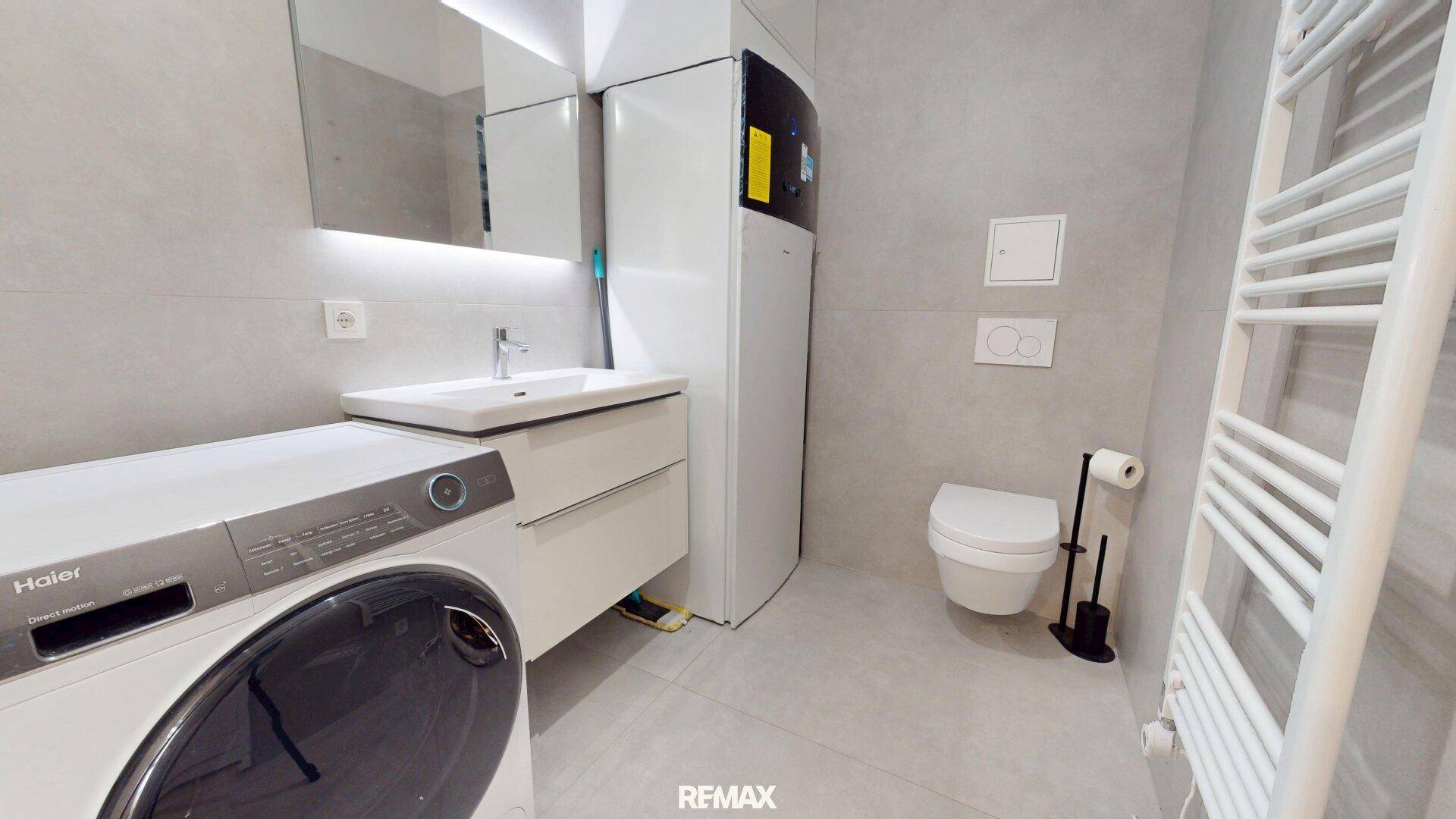 Modernes Badezimmer mit grauen Fliesen, Waschbecken, Toilette und Waschmaschine.
