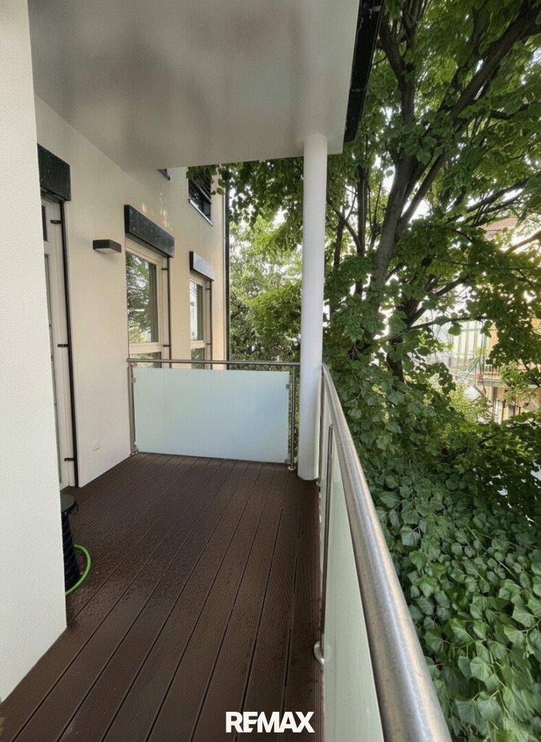 Moderner Balkon mit Holzboden und Glasgeländer, umgeben von üppigem Grün.