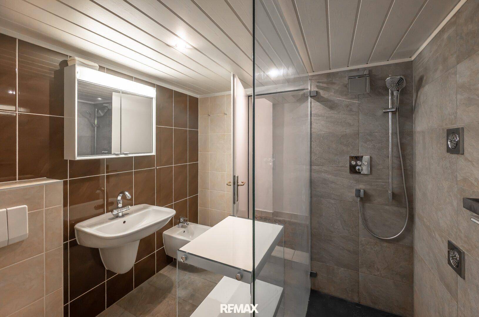 Modernes Badezimmer mit Duschkabine, Bidet, Waschbecken und braunen Fliesen.