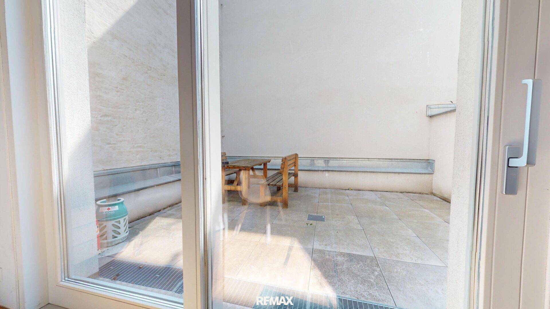 Kleine, geflieste Terrasse mit Holztisch und Stühlen, umgeben von hohen Wänden.