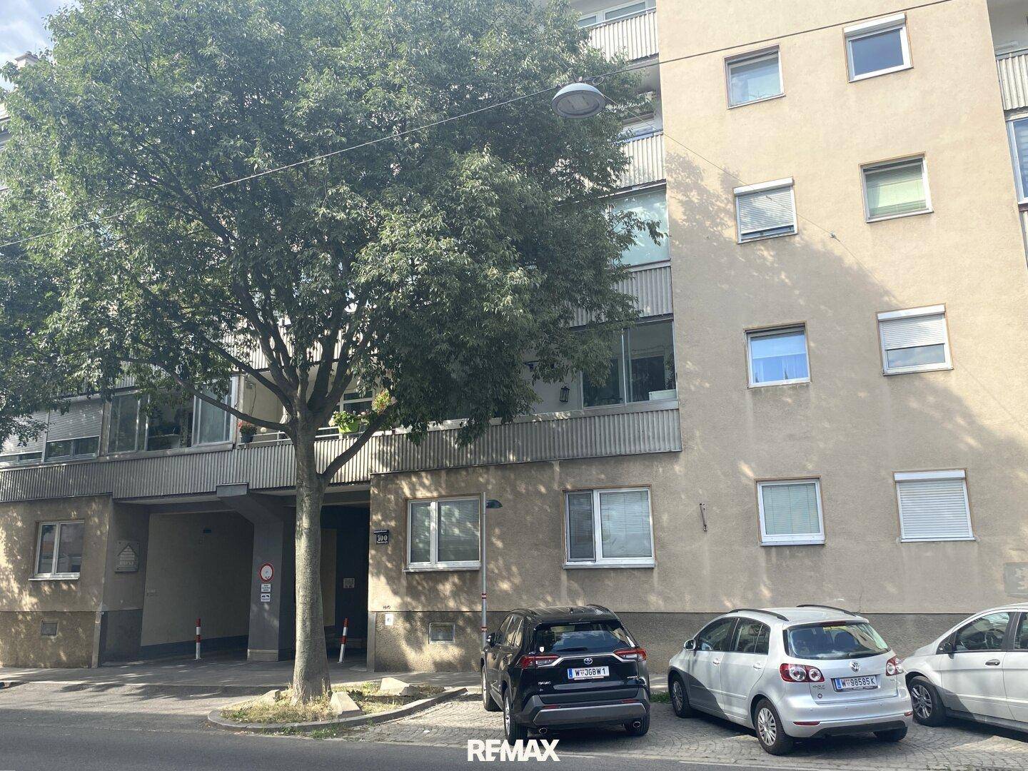 Fassade eines Mehrfamilienhauses mit geparkten Autos und Bäumen davor.