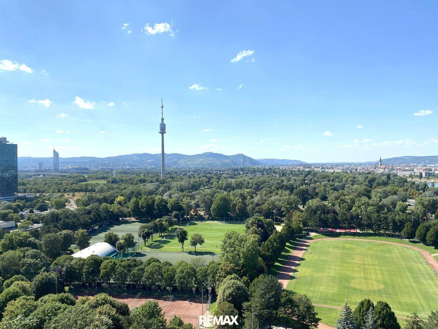 Panoramablick über die Stadt mit weiten Grünflächen, Sportplätzen und Bergen im Hintergrund.