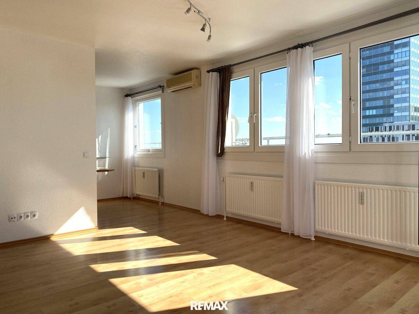 Geräumiges Wohnzimmer mit Holzboden, großen Fenstern und Klimaanlage für angenehmes Raumklima.