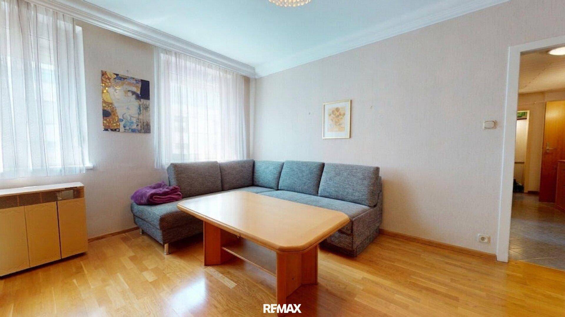 Wohnzimmer mit Ecksofa, Couchtisch und Parkettboden, hell durch große Fenster.