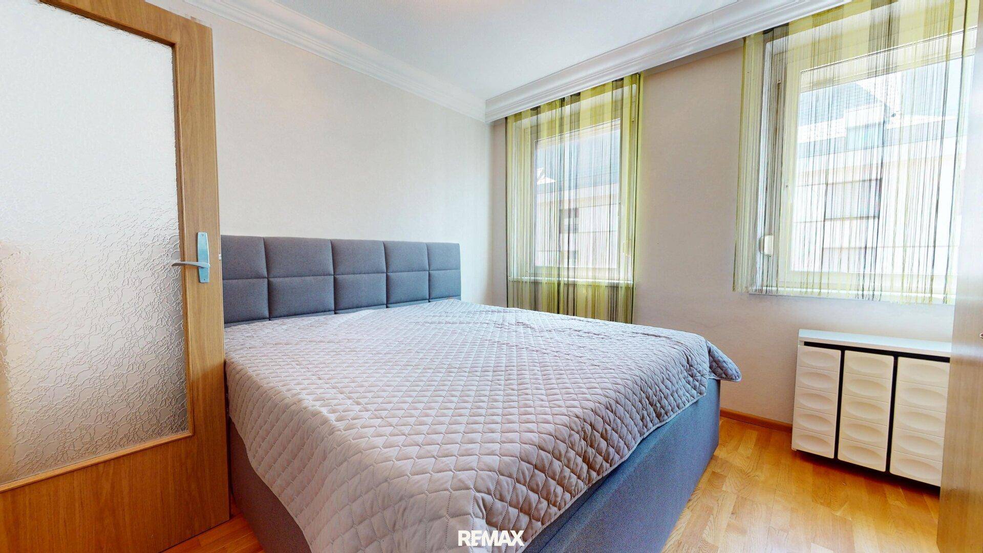 Schlafzimmer mit Doppelbett, Parkettboden und Fenstern mit hellgrünen Vorhängen.