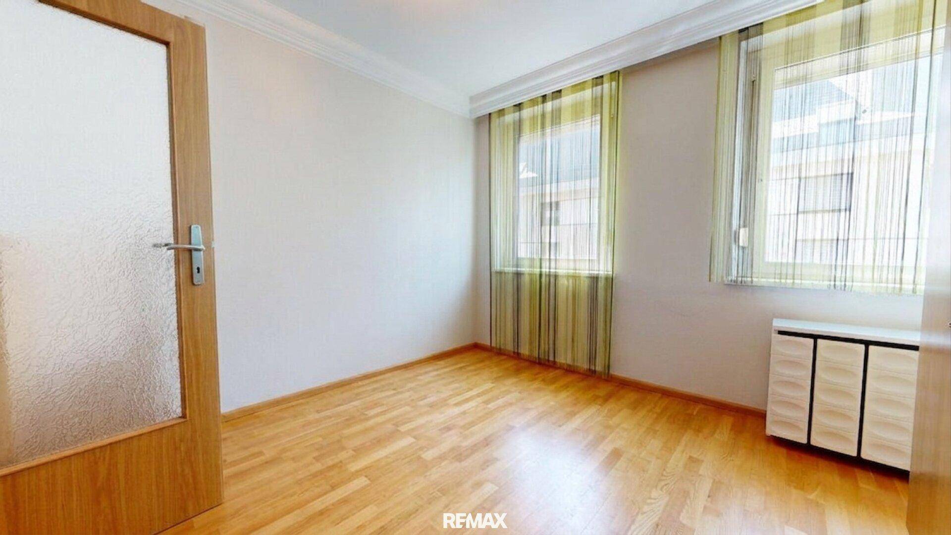 Helles, leeres Schlafzimmer mit Parkettboden und Fenstern mit gestreiften Vorhängen.