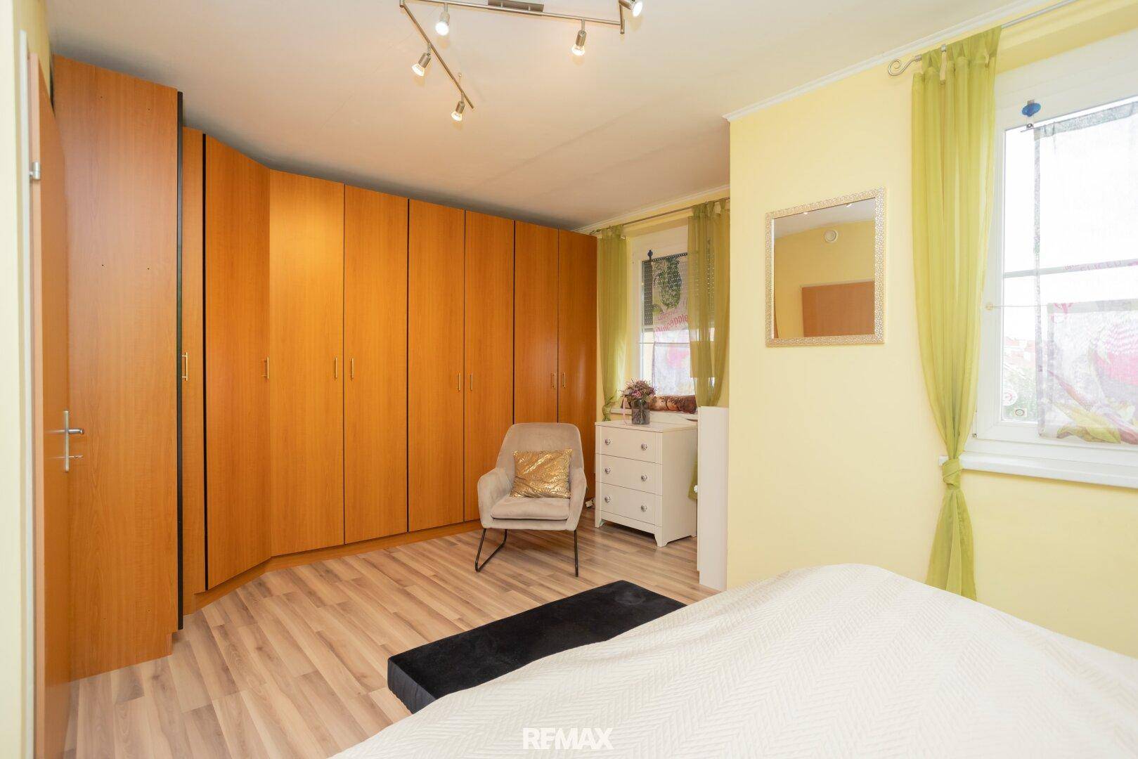 Schlafzimmer mit großen Holzschränken, Laminatboden und einem Fenster mit gelben Vorhängen.