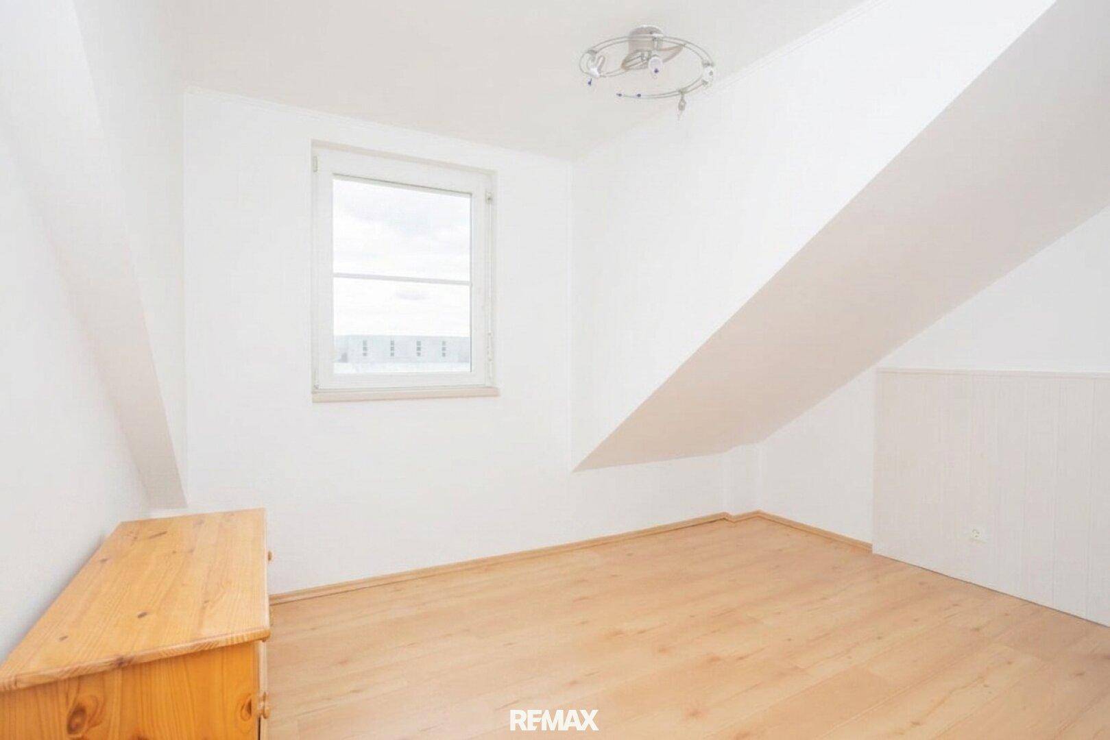 Leerer Raum mit Fenster, schräger Decke und hellem Laminatboden, vielseitig nutzbar.