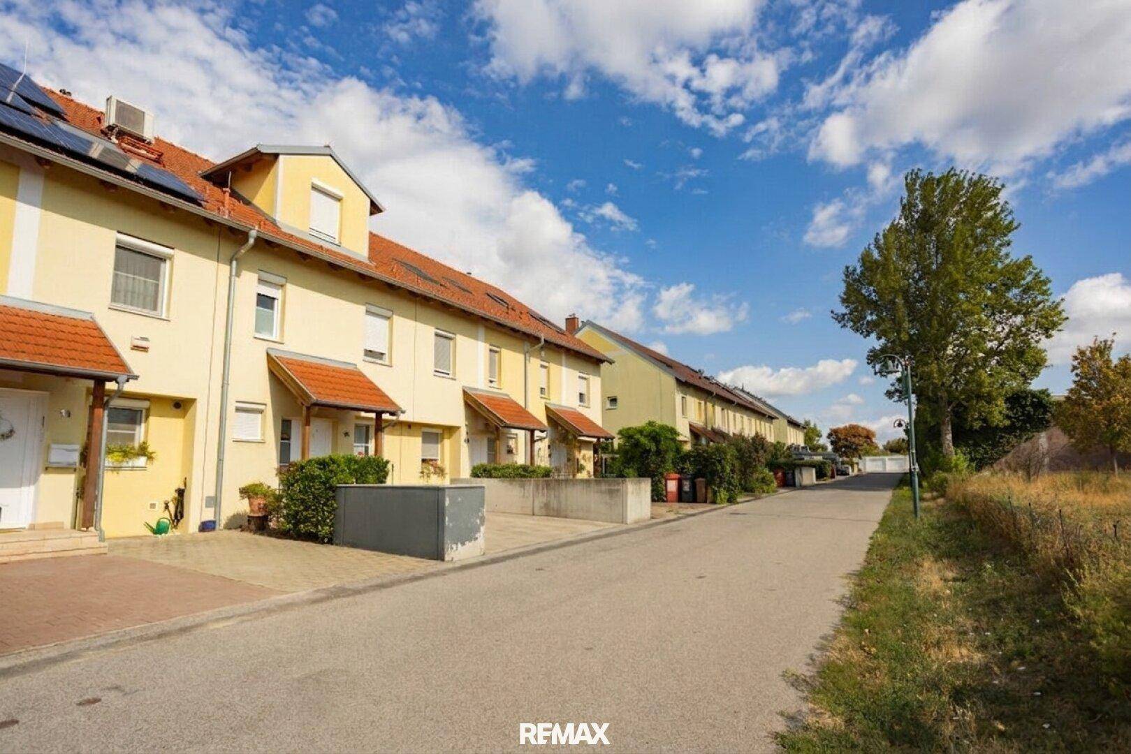 Reihenhäuser entlang einer Straße mit Vorgärten und Zufahrten unter blauem Himmel.