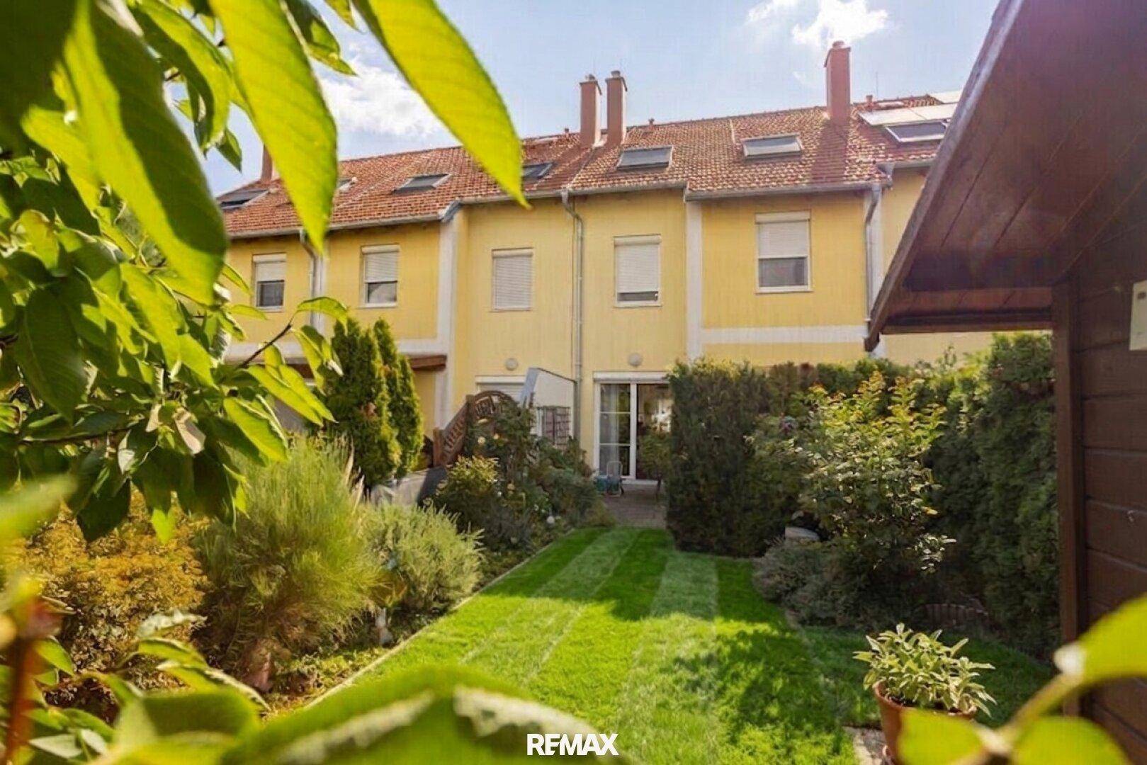Gepflegter Gartenbereich mit Rasen und üppiger Bepflanzung hinter einer Reihe von Wohnhäusern.