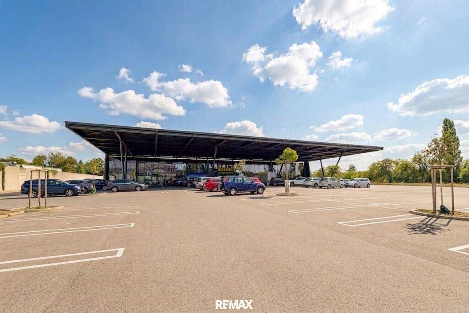 Modernes Geschäftsgebäude mit weitläufigem Parkplatz und markanter Dachkonstruktion unter blauem Himmel.