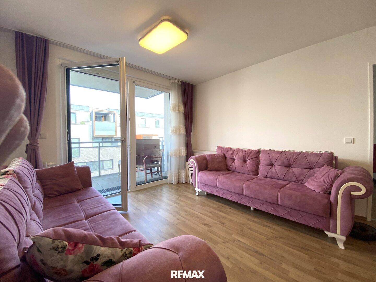 Wohnzimmer mit Zugang zum Balkon, Holzboden und lila Vorhängen.