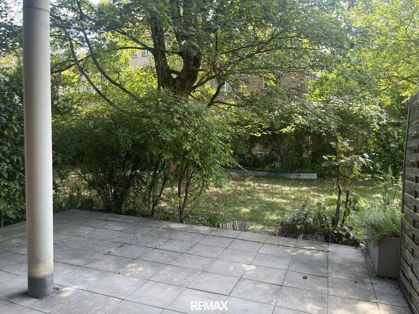 Helle Terrasse mit Steinplatten und üppigem Grün, das den Garten umgibt.